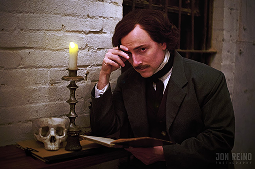 Campbell Harmon as Edgar Allan Poe, leaned over a small desk against a brick wall, holding a thin folio in one hand. The other hand against his forhead, lightly holding a pen. A skull and candle are on the desk.